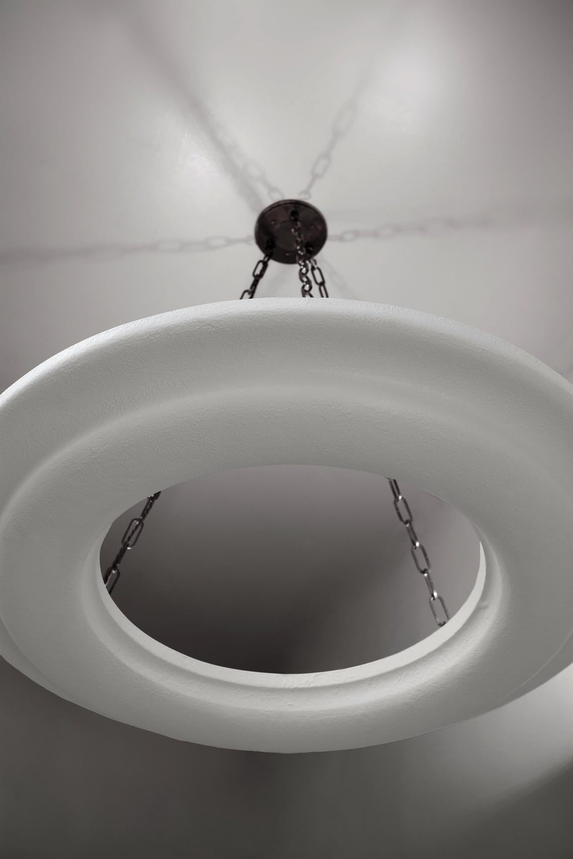 An angled shot looking up at a large, circular white chandelier with a smooth, matte finish. It is suspended from a light-colored ceiling by three dark chains converging at a central ceiling plate, casting subtle shadows that fan out around it.