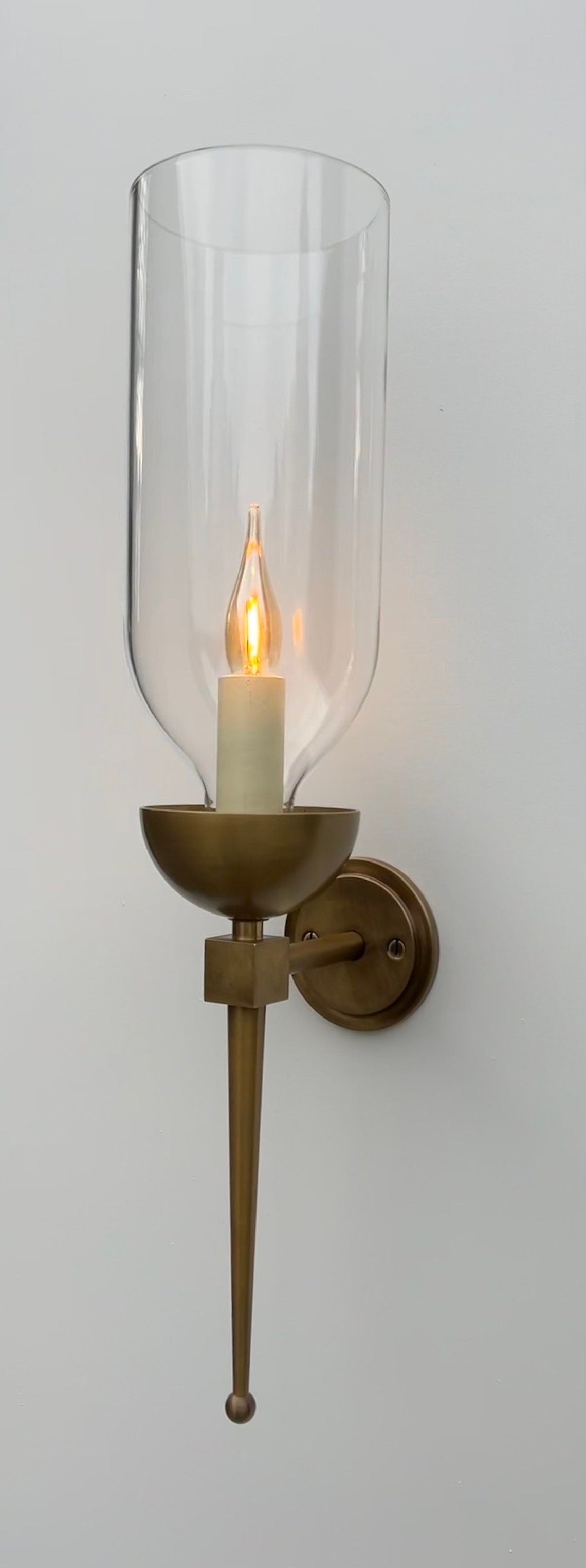 Brass wall sconce with an illuminated candle bulb and clear glass hurricane.