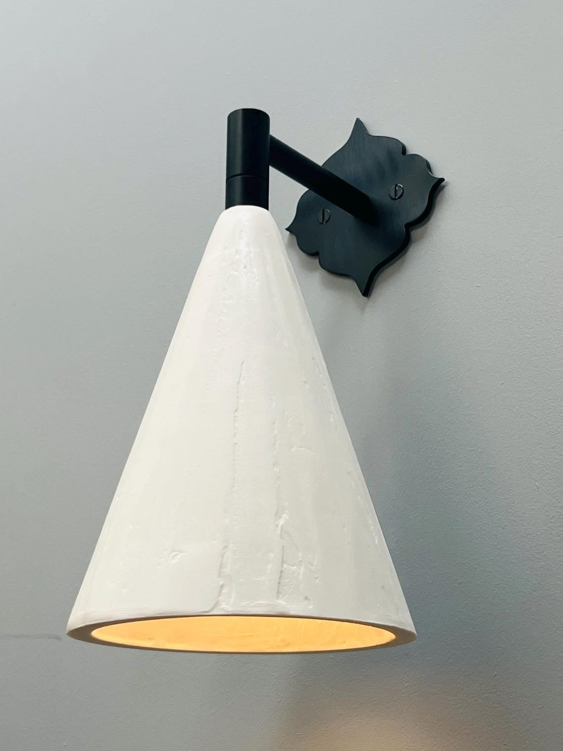 A wall-mounted sconce with a black, ornate, scroll-shaped backplate and a short, black arm holding a textured, off-white conical shade. Warm yellow light emanates from the bottom of the shade, casting a soft glow on the light gray wall beneath it.
