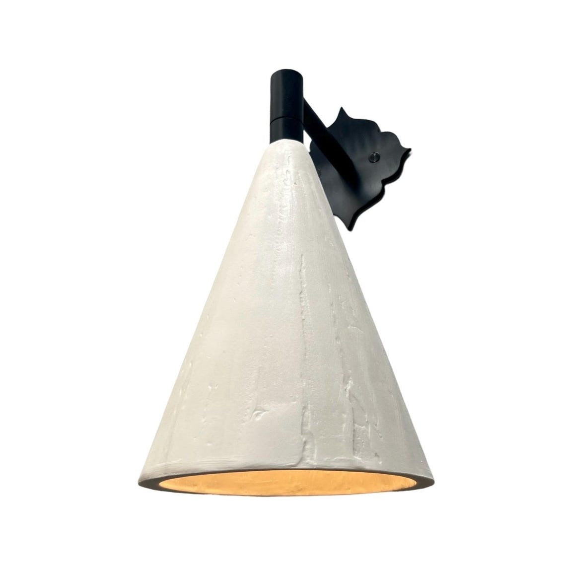 A wall-mounted sconce with a matte black, ornate, scroll-shaped backplate and a short, black arm, holding a downward-facing, textured off-white conical shade. A warm, yellow-orange light glows from the interior of the shade, all set against a plain white background.