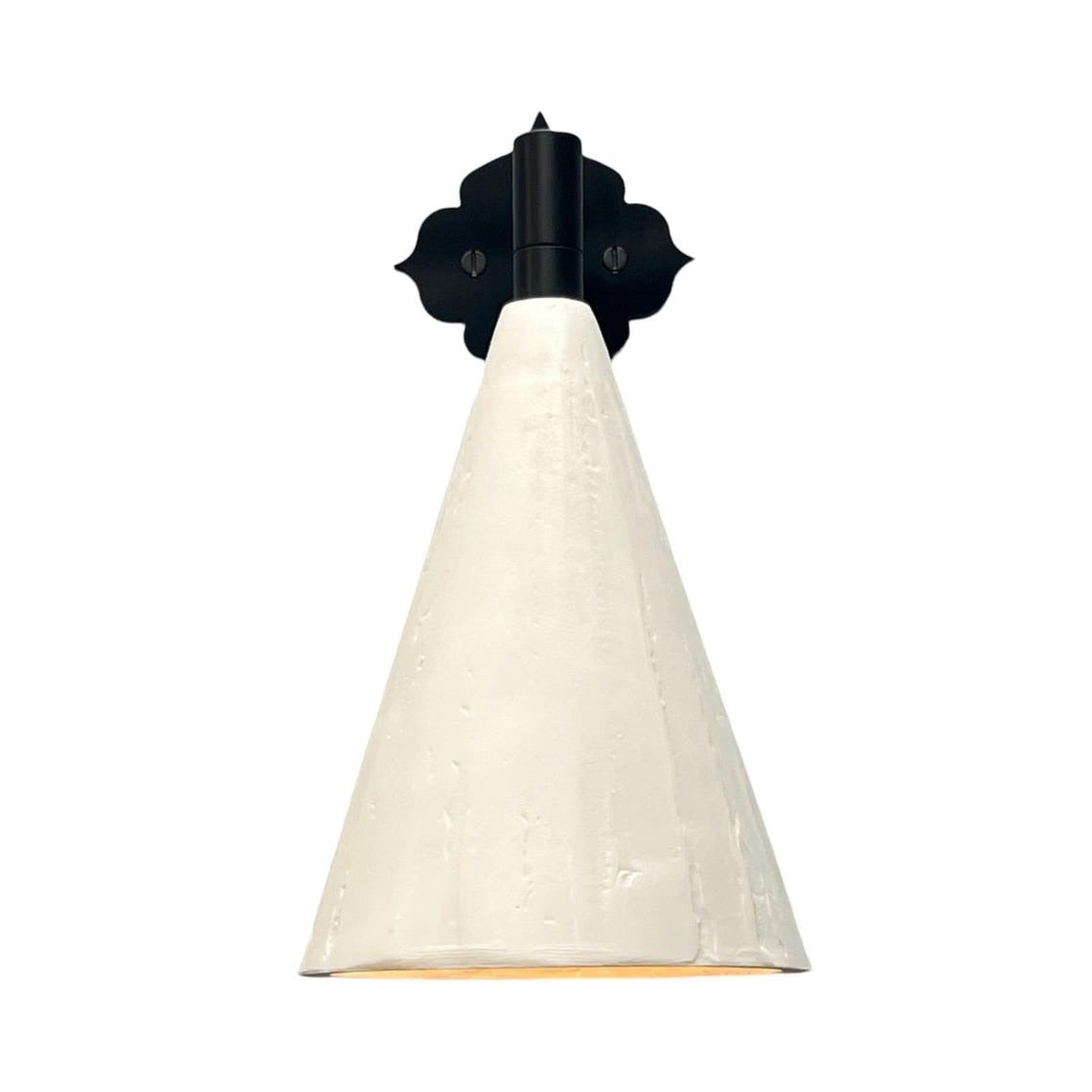 A wall-mounted sconce with a matte black, ornate, scroll-shaped backplate and a short, black arm, holding a downward-facing, textured off-white conical shade. A warm, yellow-orange light glows from the interior of the shade.