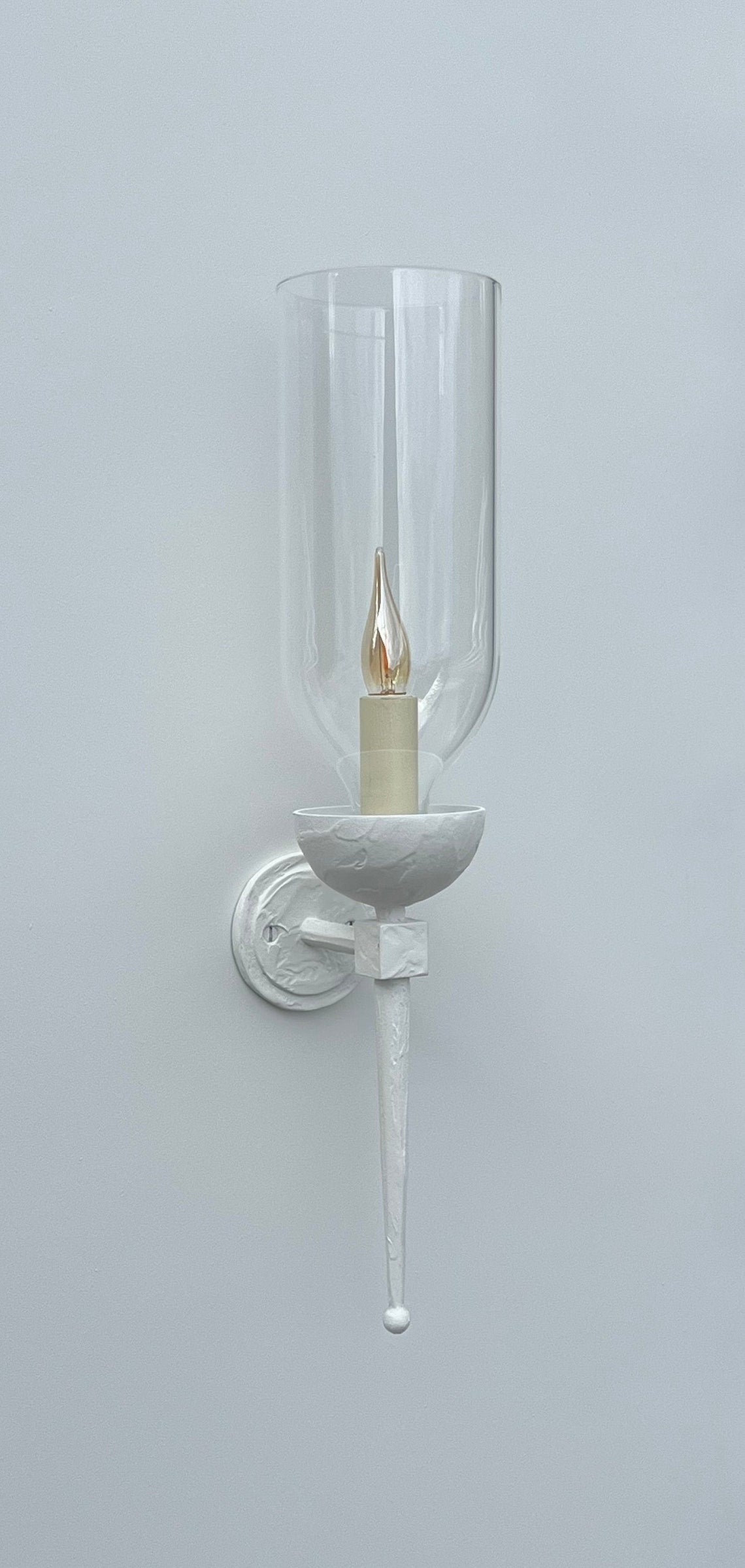White wall sconce with clear glass and a flickering candle-style bulb.