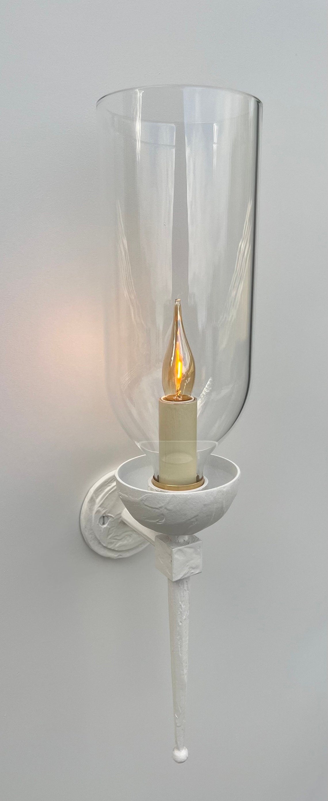 Side view of a white wall-mounted sconce. The fixture features a circular backplate, an arm, and a bowl-shaped holder, all in white. A clear, tall glass hurricane shade encloses a glowing, flickering candle-style light bulb. A long, tapered finial extends downwards from the base of the fixture.