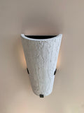 A tall, inverted cone-shaped wall sconce with a textured, bark-like white finish. It is mounted on a light-colored wall with three small black square brackets visible