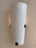 A tall, inverted cone-shaped wall sconce with a textured, bark-like white finish. It is mounted on a light-colored wall with three small black square brackets visible.