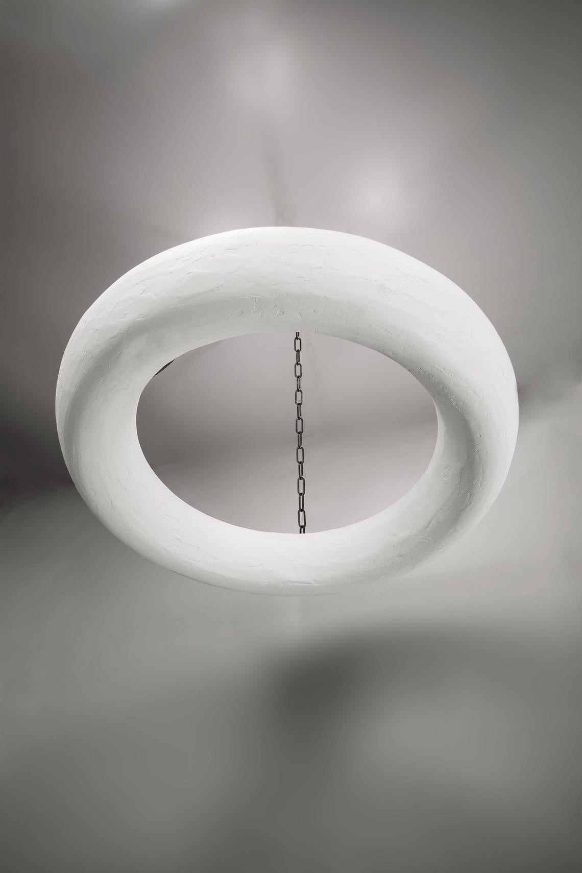  A modern, off-white, textured circular chandelier is suspended from a dark, round ceiling plate by four dark metal chains. The light fixture casts subtle shadows on the plain gray ceiling above.