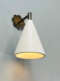  A modern wall sconce with a white conical shade and antique brass floral backplate, illuminated against a gray wall.