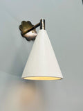  A modern wall sconce with a white conical shade and antique brass floral backplate, illuminated against a gray wall.