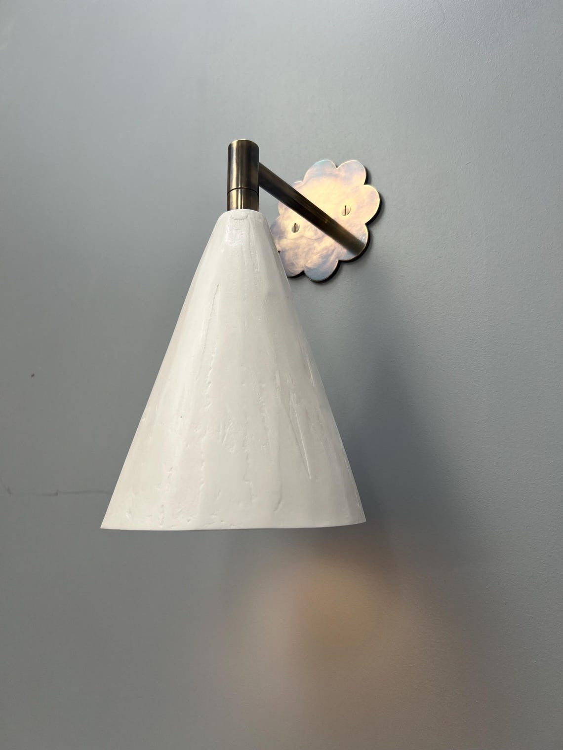 A white conical wall sconce with a floral brass backplate on a gray wall.