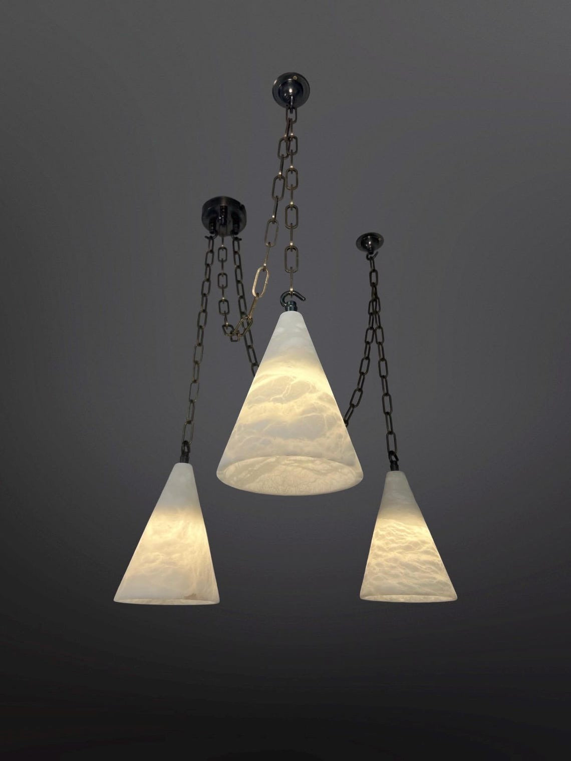 Three pendant lights with textured, translucent white conical glass shades hang at varying heights from a dark ceiling. Each light is suspended by a dark metal chain, and the shades are illuminated from within, showcasing their swirled or rippled patterns against a dark gray background.