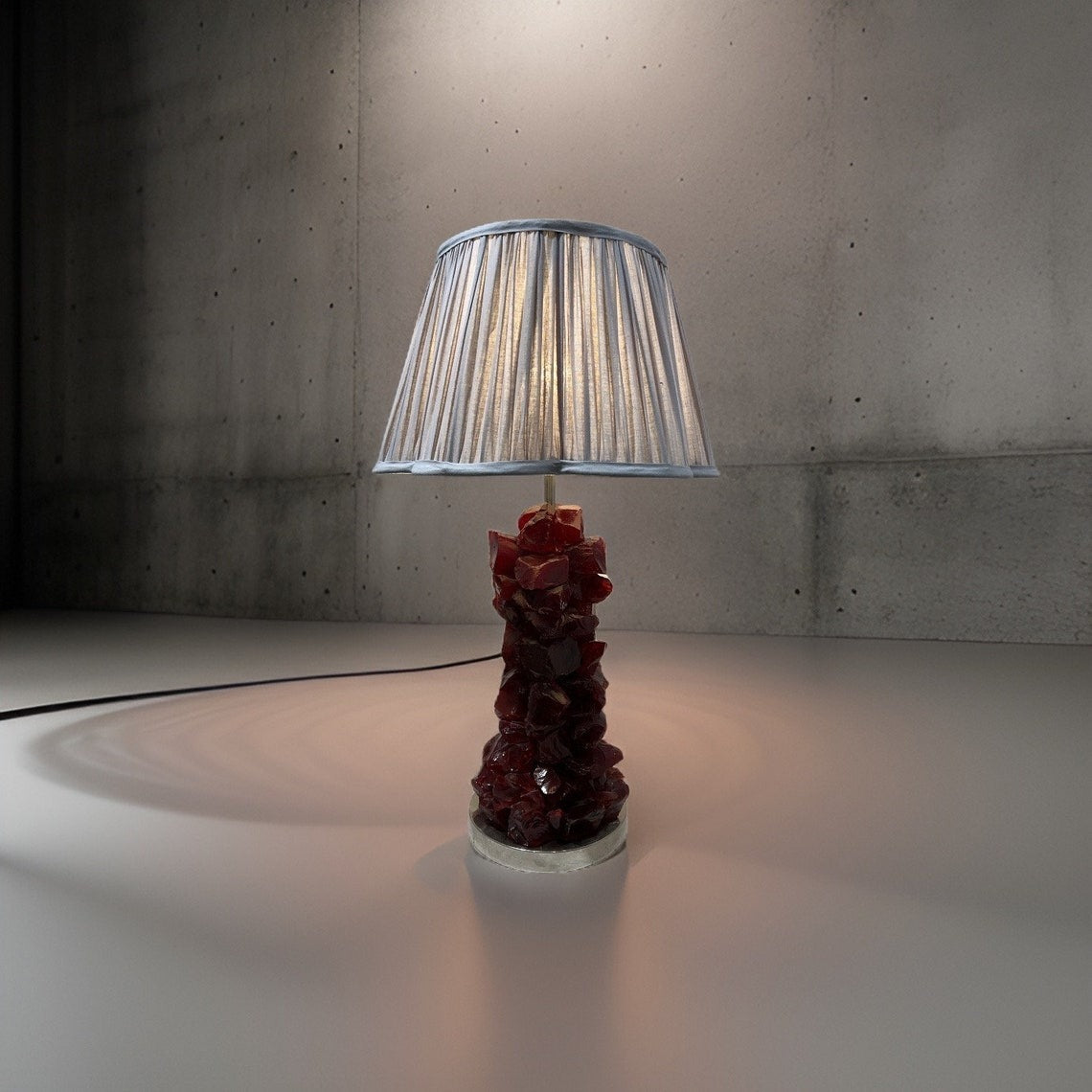 A table lamp with a dark red, textured base and a pleated light-colored shade, casting warm light on a concrete floor and wall.