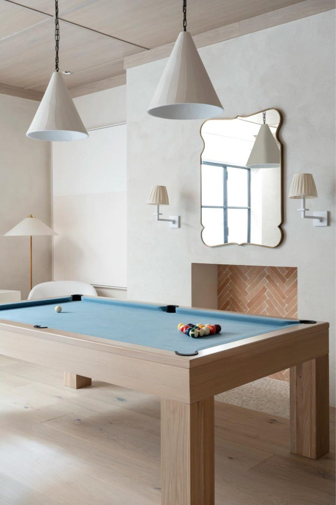 A modern light-wood pool table with a light blue felt surface is centered in a room with two large conical pendant lights, a fireplace, and a mirror.