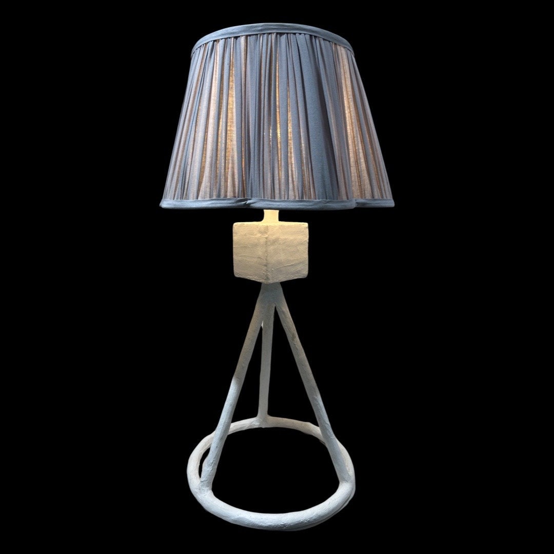 A tall white table lamp with a tripod base and pleated grey lampshade on a black background.