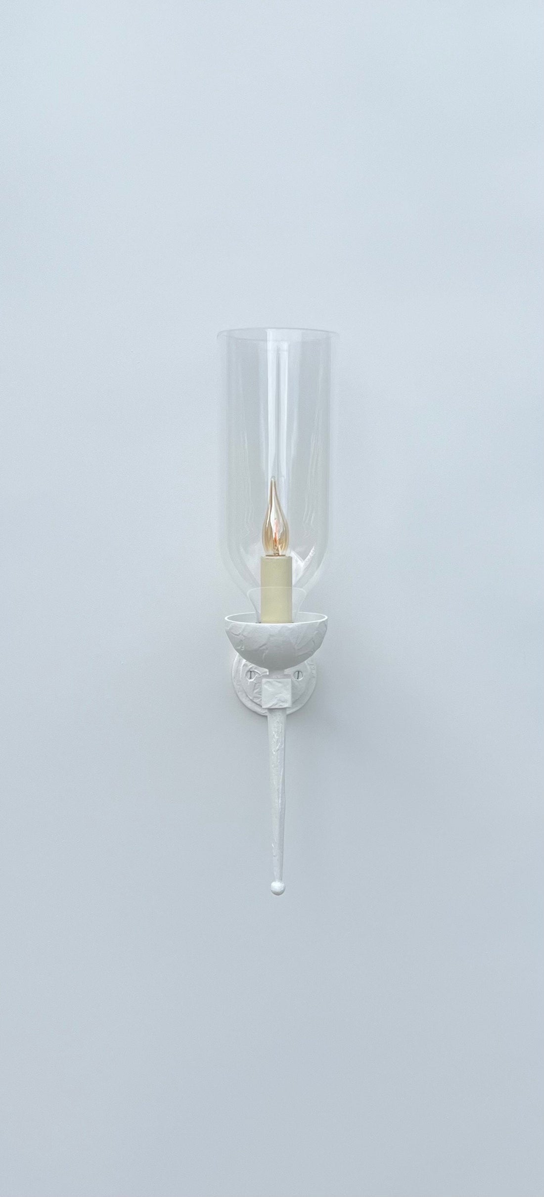
A front-on view of a white wall-mounted sconce against a plain white wall. The fixture features a circular backplate, an arm, and a bowl-shaped holder, all in white. A clear, tall glass hurricane shade encloses a glowing, flickering candle-style light bulb. A long, tapered finial extends downwards from the base of the fixture.