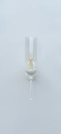 
A front-on view of a white wall-mounted sconce against a plain white wall. The fixture features a circular backplate, an arm, and a bowl-shaped holder, all in white. A clear, tall glass hurricane shade encloses a glowing, flickering candle-style light bulb. A long, tapered finial extends downwards from the base of the fixture.