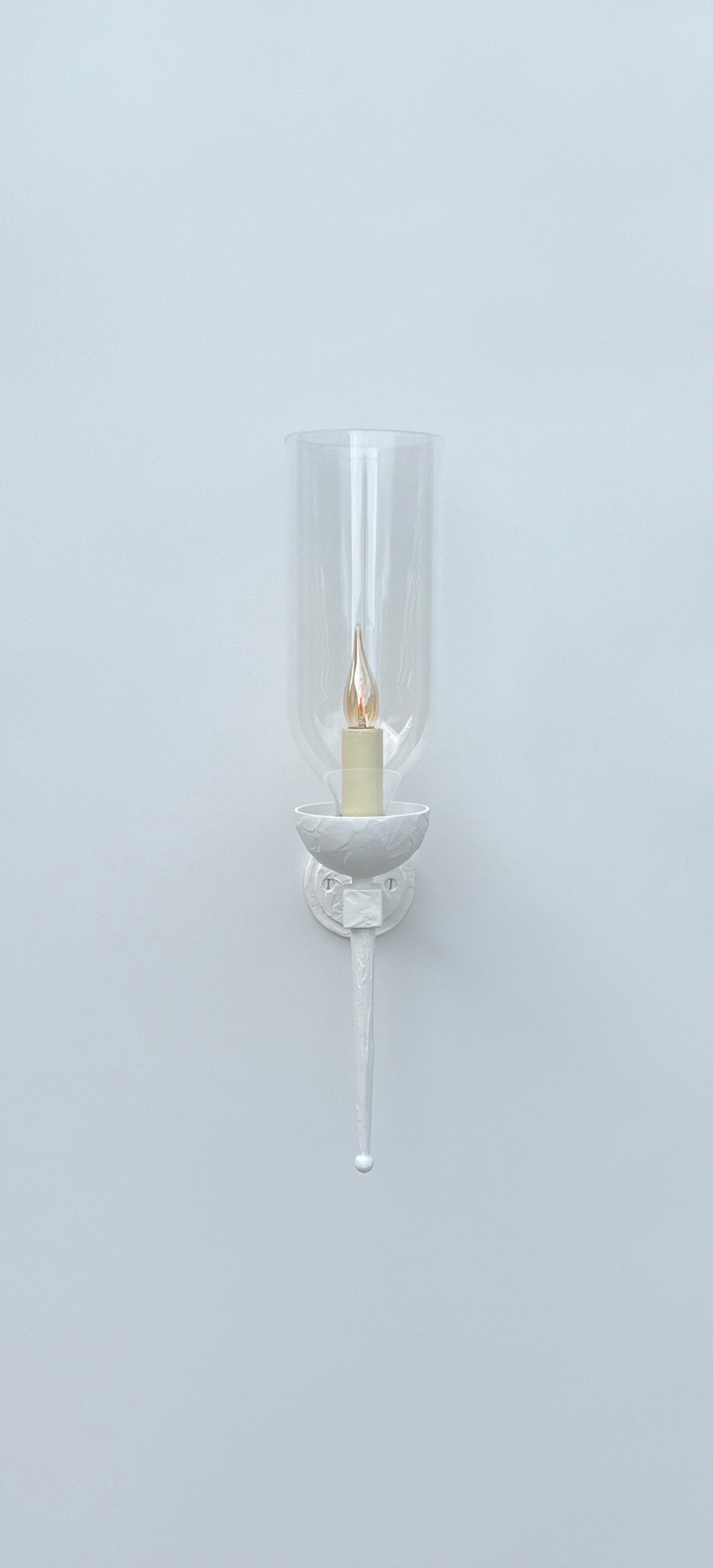 
A front-on view of a white wall-mounted sconce against a plain white wall. The fixture features a circular backplate, an arm, and a bowl-shaped holder, all in white. A clear, tall glass hurricane shade encloses a glowing, flickering candle-style light bulb. A long, tapered finial extends downwards from the base of the fixture.