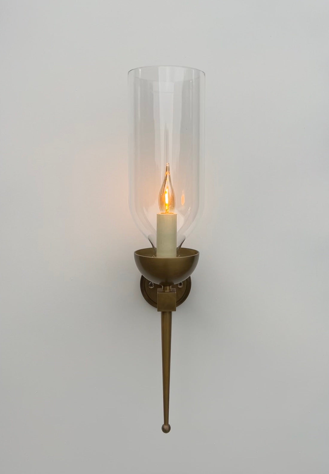 A front-on view of a wall-mounted sconce against a plain white wall. The fixture features a circular backplate, an arm, and a bowl-shaped holder, all with a textured, patinated green (verdigris) copper finish. A clear, tall glass hurricane shade encloses a glowing, flickering candle-style light bulb. A long, tapered finial extends downwards from the base of the fixture.