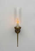A front-on view of a wall-mounted sconce against a plain white wall. The fixture features a circular backplate, an arm, and a bowl-shaped holder, all with a textured, patinated green (verdigris) copper finish. A clear, tall glass hurricane shade encloses a glowing, flickering candle-style light bulb. A long, tapered finial extends downwards from the base of the fixture.