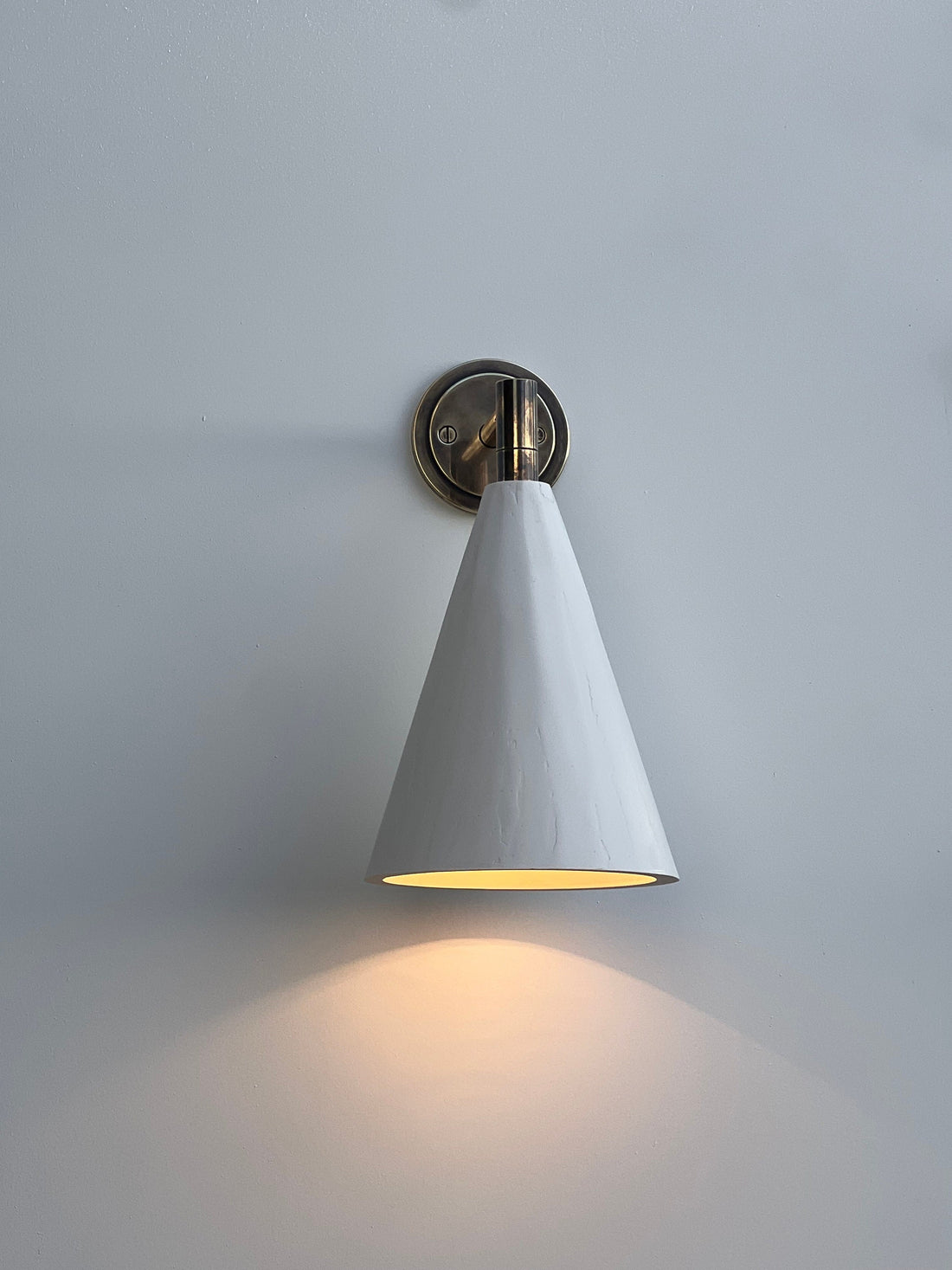 A modern brass and plaster cone wall light with a bronze base on a white wall, casting a warm glow downwards.