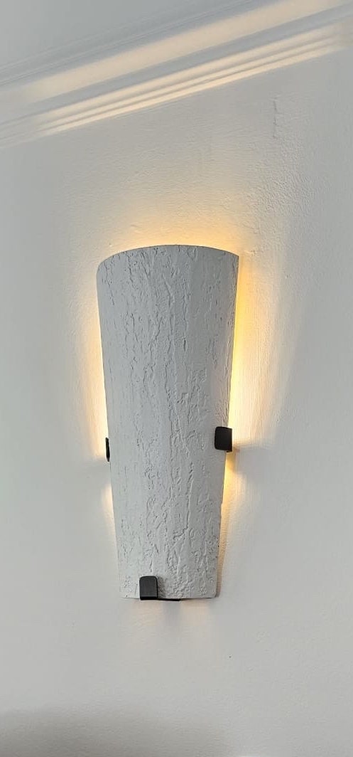 A modern, textured white wall sconce casting warm light upwards and downwards against a light-colored wall. It's held by dark, square brackets.