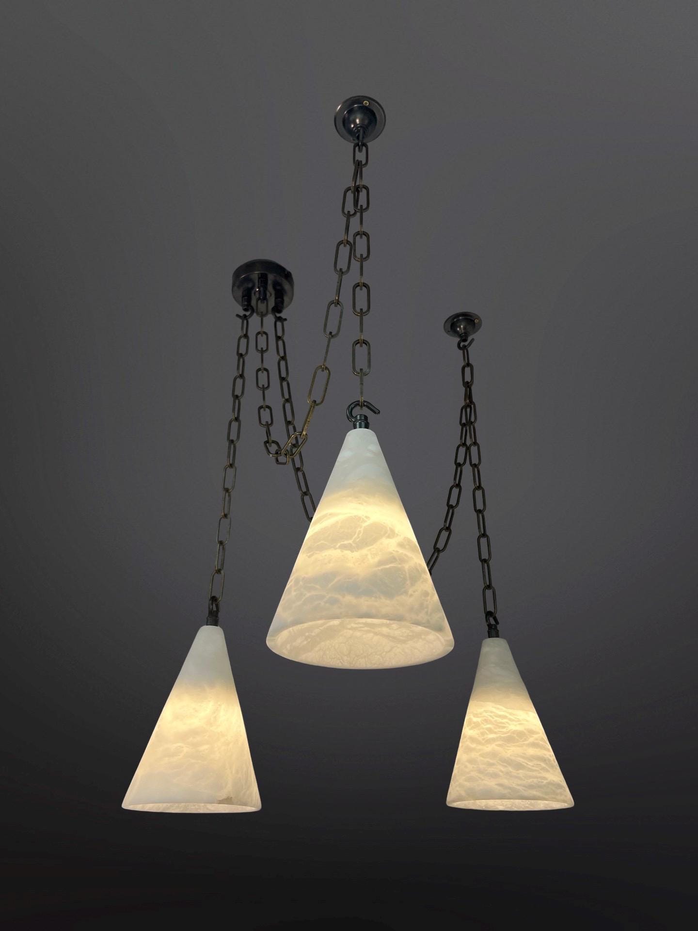 Three pendant lights with textured, translucent white conical glass shades hang at varying heights from a dark ceiling. Each light is suspended by a dark metal chain, and the shades are illuminated from within, showcasing their swirled or rippled patterns against a dark gray background.