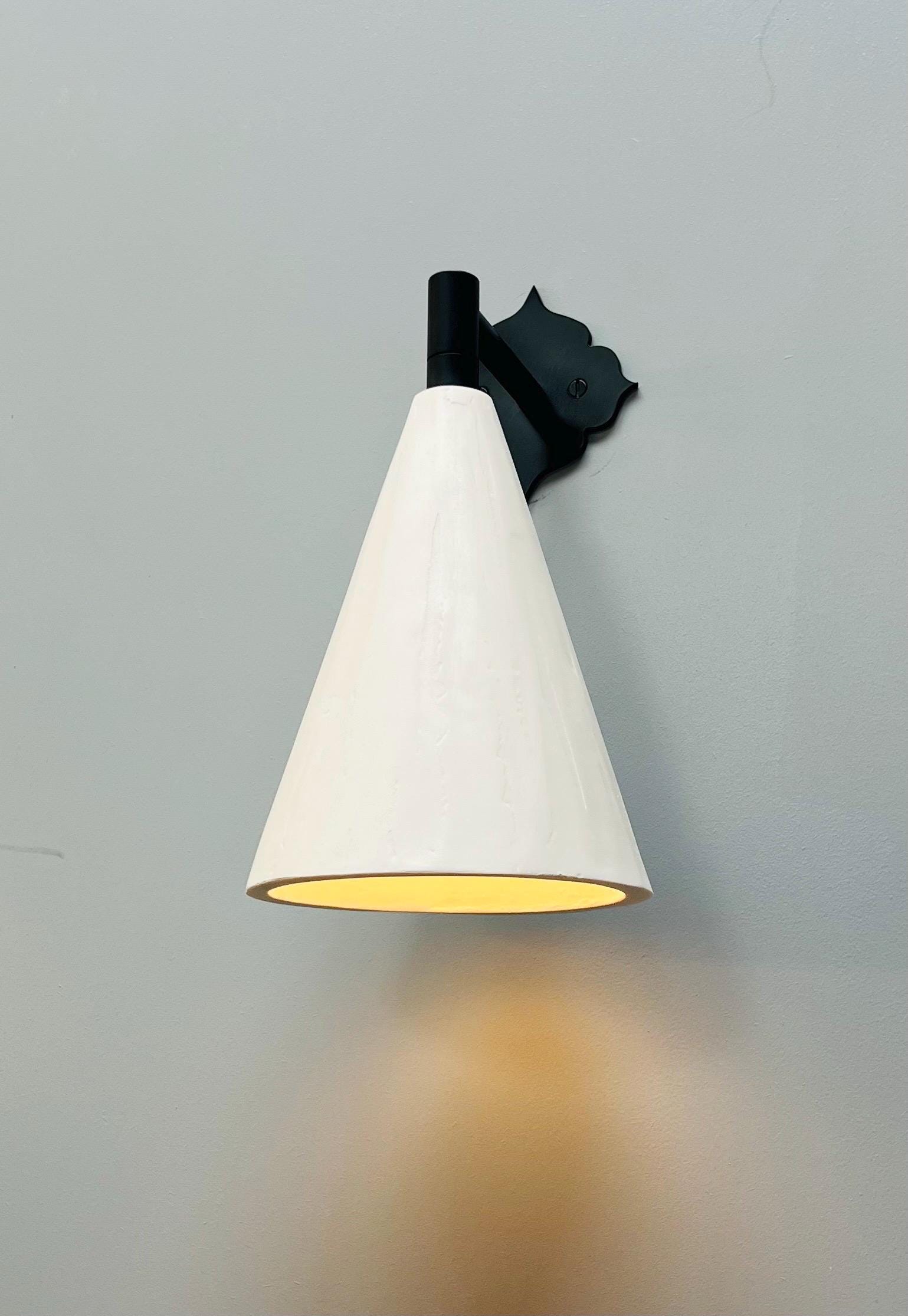 White conical wall sconce with a decorative black backplate, casting warm light.