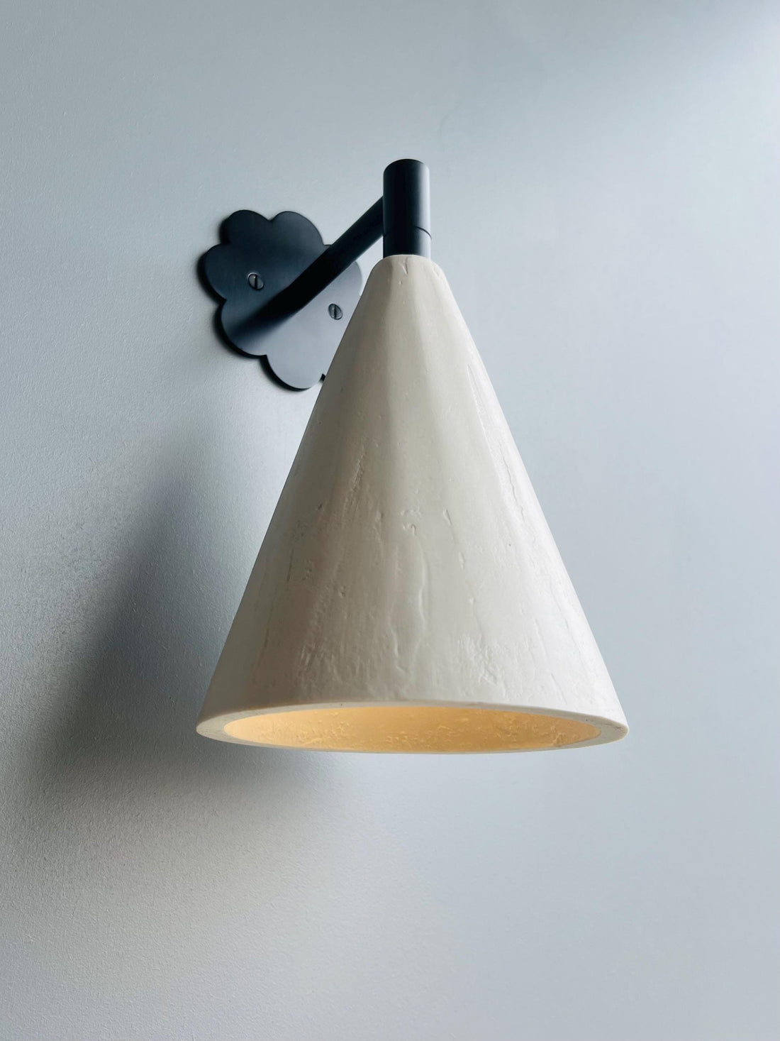A modern wall sconce with a light conical shade and dark floral backplate, illuminated against a light gray wall.